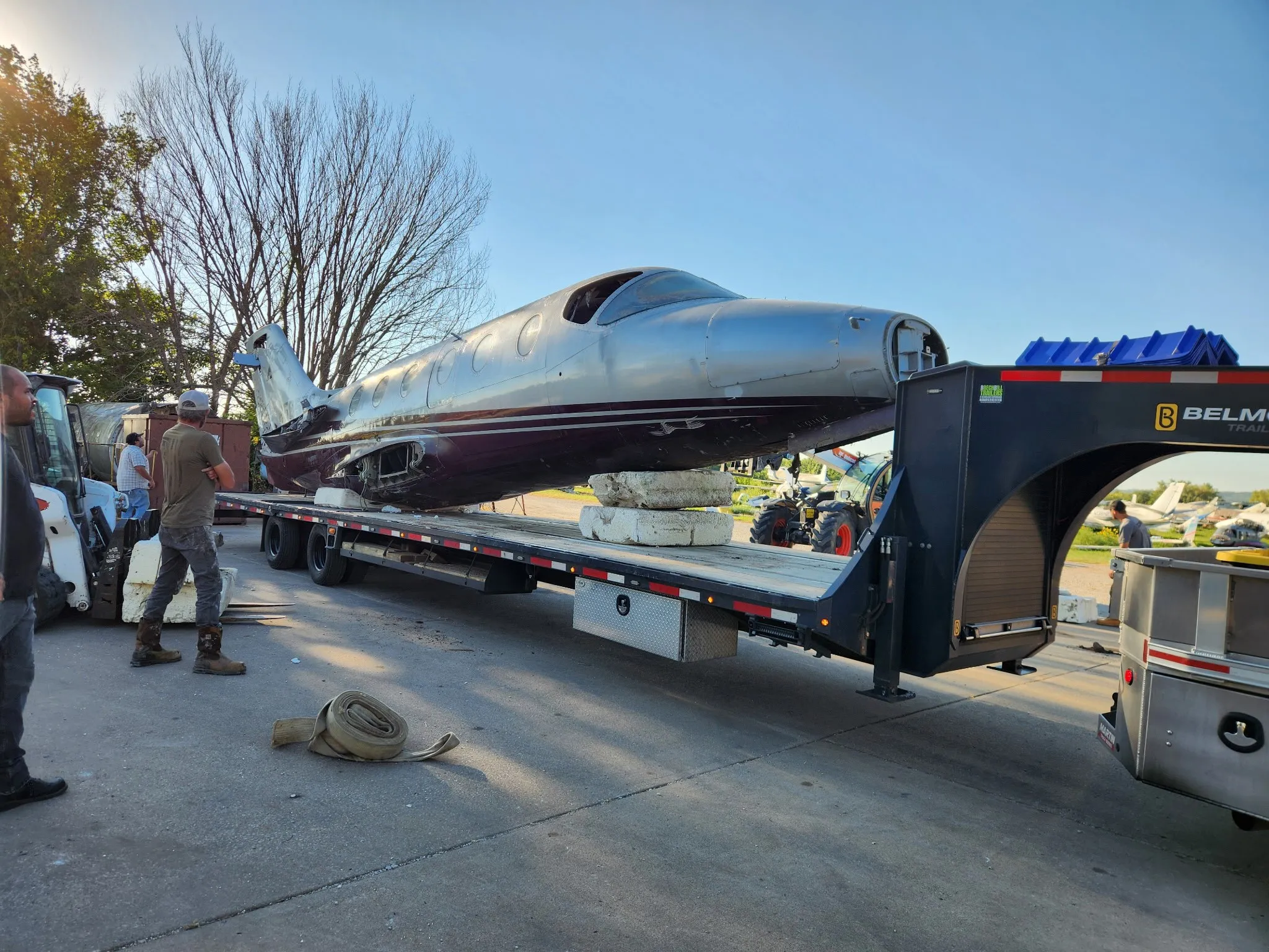 The aircraft fuselage on a flatbed trailer, with people and lifting equipment nearby.