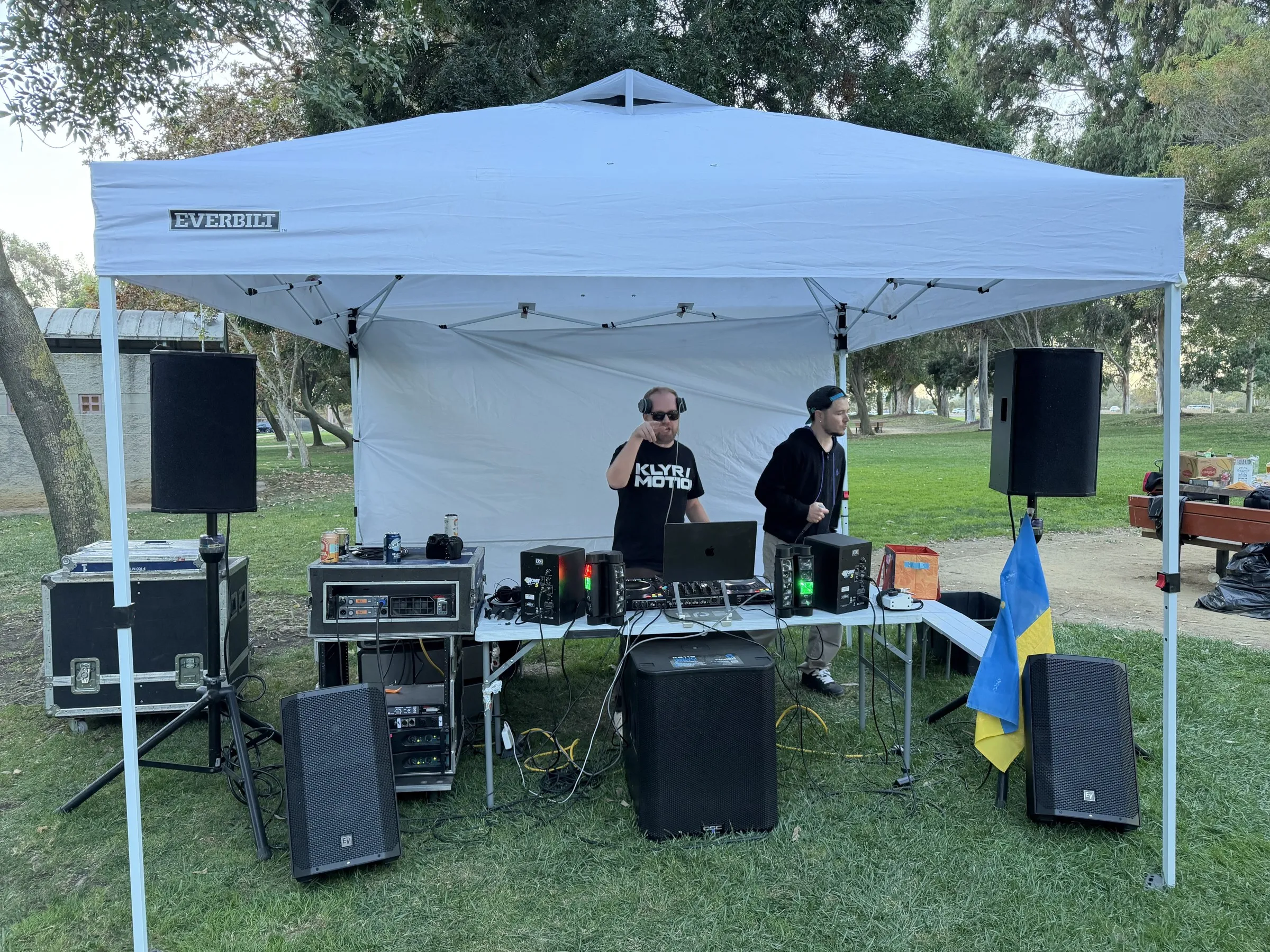 Two DJs at an outdoor setup beneath a white canopy, with a Ukrainian flag beside the speakers.